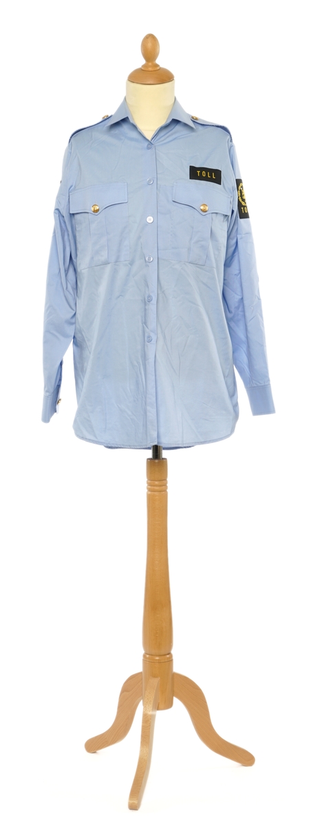 Lys blå langermet skjorte av et fintvevd stoff. Epauletter uten tilsatt distinktsjon. Tekstil merke på begge armer: riksløve med hvit øks omgitt av eikeblader over TOLL. Tekstil merke på venstre bryst, gul bokstaver på sort, 'TOLL'.