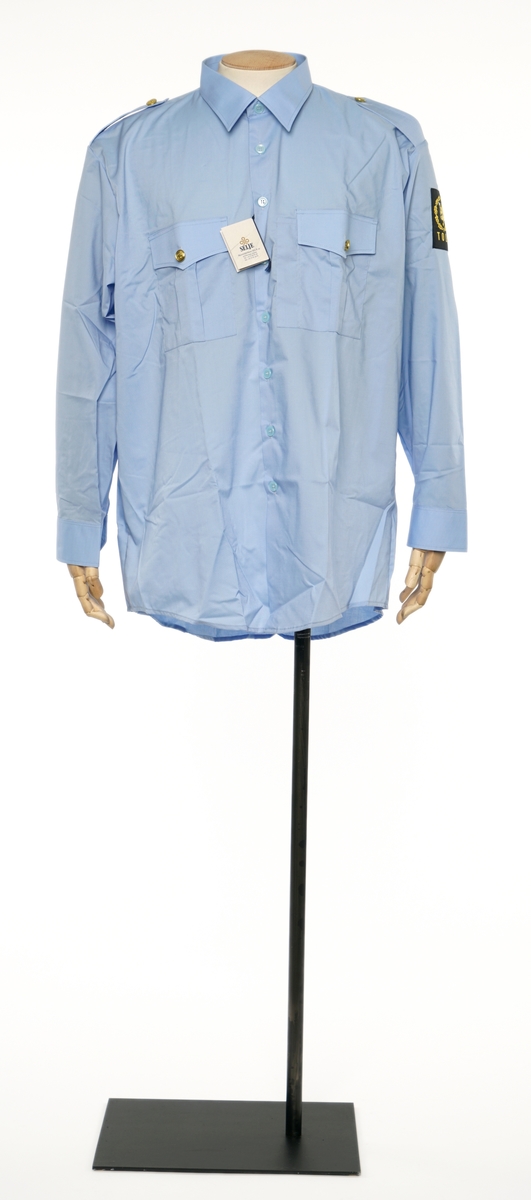 Lys blå  langermet skjorte av et fintvevd stoff. Epauletter uten tilsatt distinktsjon. Tekstil merke på venstre armen: riksløve med hvit øks omgitt av eikeblader over TOLL.