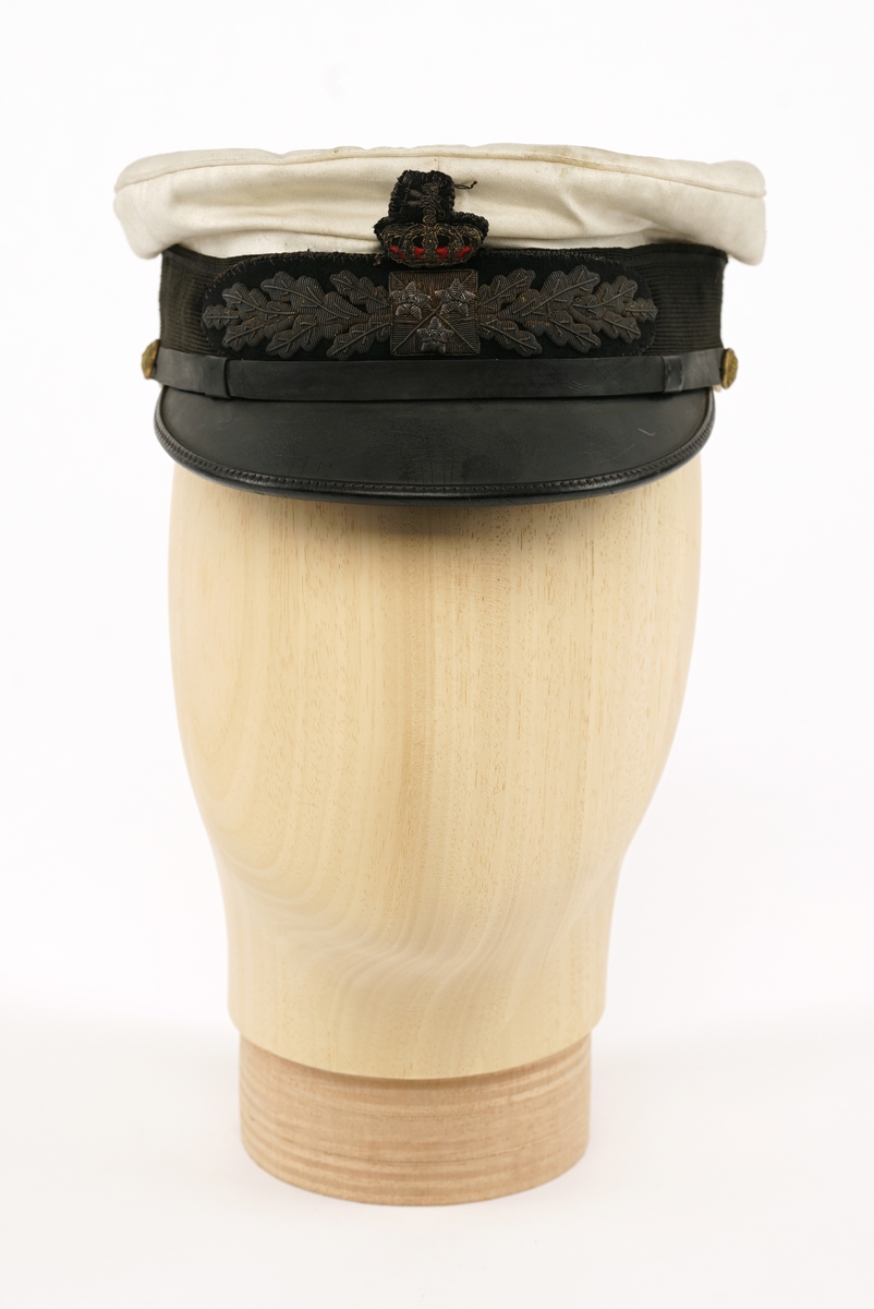 Hvit hatt med sort visir. Sort plast/lær strimmel/bånd over visir festet med riksløvesknapper på hver side. Merke på framsiden, krone over to eikekvister med tre stjerner i firkant.