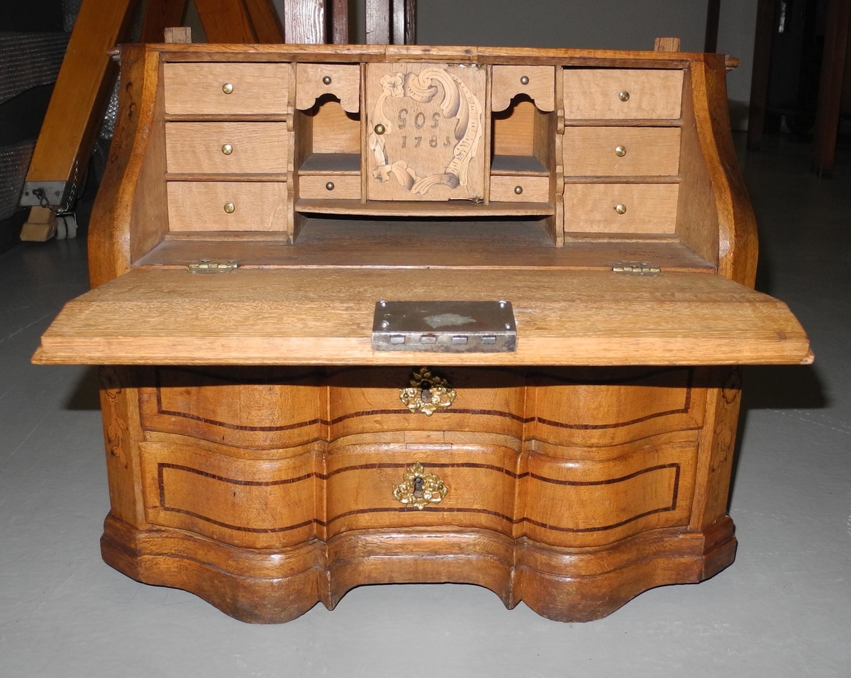 Barneskatoll i rokokkostil med separat overskap og underskap samt felles nøkkel til begge deler. Underskap med kraftig buet og profilert front, to skuffer samt nedfellbart pultlokk med hylle-, skap- og skuffseksjon innenfor. Overskap med to rette dører og kraftig profilert, delvis gjennombrutt gesims. Både over- og underskapet rikt dekorert med intarsia av høy kvalitet med rocailler, skjell, konturlinjer og blomsterornamentikk. Dekorative skilt av messing rundt alle nøkkelhull. Initialer og årstall på skapdør bak pultlokk. Skatollet står på to lave føtter.

A: underskap
B: nøkkel
C: overskap