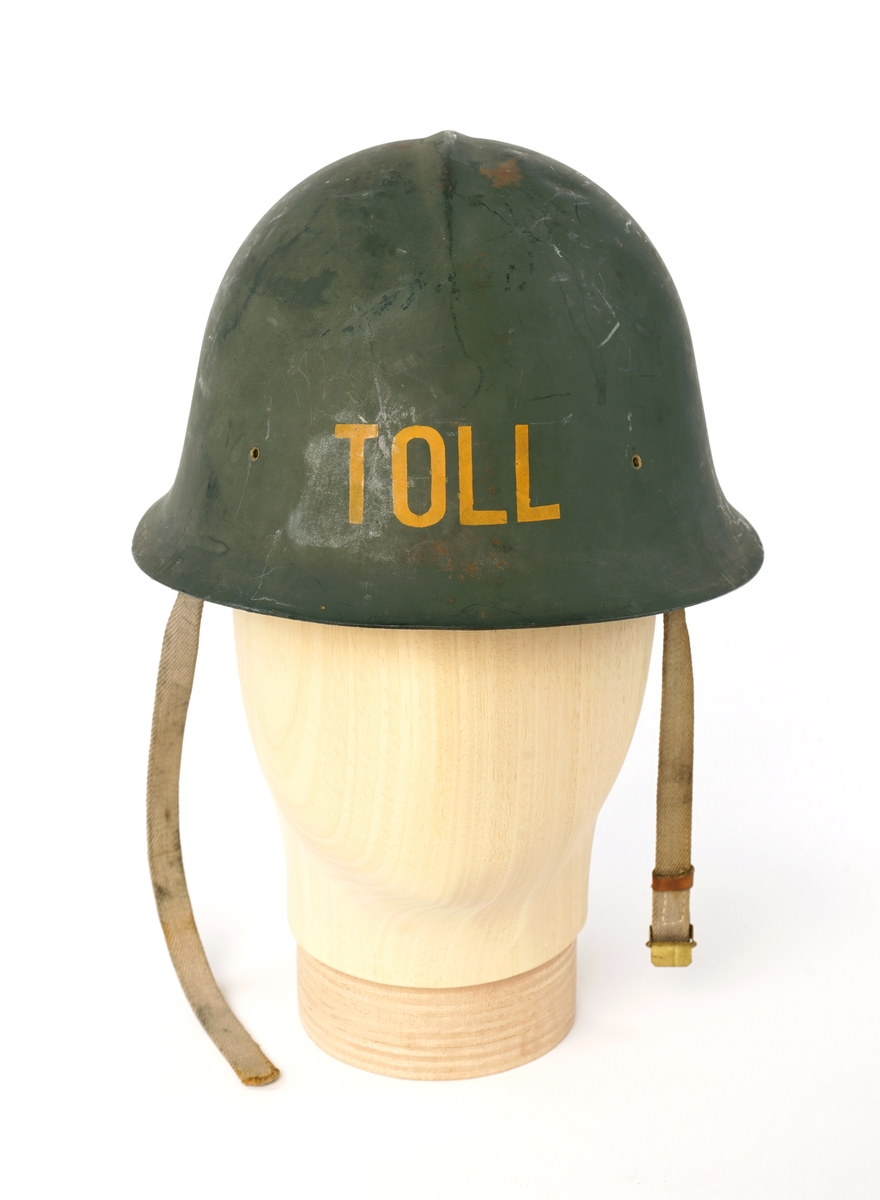 Metall hjelm (magnetisk), grønnmalt med 'TOLL'  stensilerte i gul maling. På innsiden, tre lær klaffer med puter på innsiden, forbundet med en sirkel av snor å justere for hodestørrelse. Tekstilbånd på hver side, en med metalllås for lukking under haken.