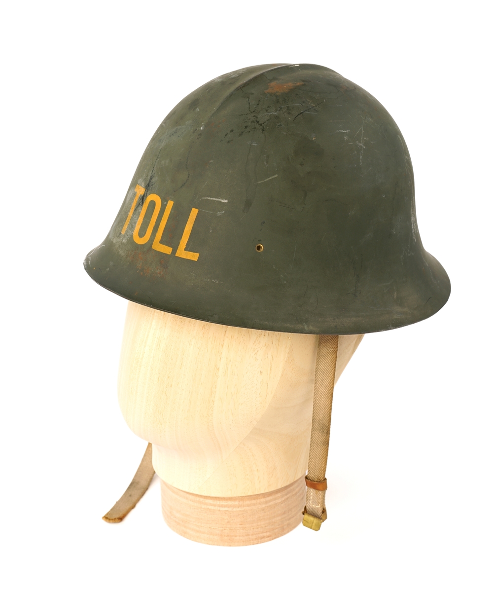 Metall hjelm (magnetisk), grønnmalt med 'TOLL'  stensilerte i gul maling. På innsiden, tre lær klaffer med puter på innsiden, forbundet med en sirkel av snor å justere for hodestørrelse. Tekstilbånd på hver side, en med metalllås for lukking under haken.