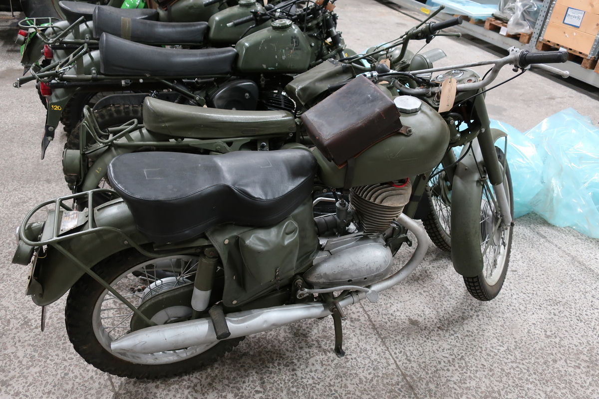 Motorcykel av märket JAWA. Hastighetsmätare graderad 0-140 km. På tanken sitter en verktygslåda av läder. Avlång sittdyna sk "limpa".