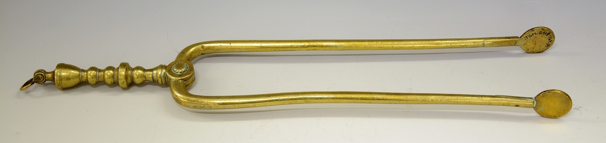 Ildtang av messing, del av peissett i tre deler. Bøyle øverst til oppheng, dreide kuler og blomst der armene deler seg. Avslutning på armene er to ovaler.