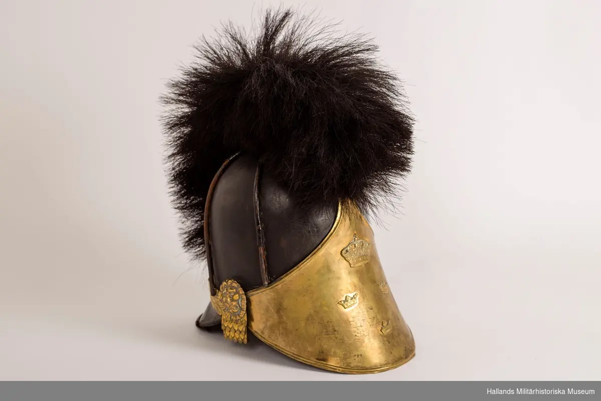 En s.k kask (hjälm) för manskap vid infanteriet. Hjälmen är gjord av styvt svart läder och har fram- och bakskärm. Framskärmens insida är mörkgrön. 
Svartlackade metalldetaljer på hjälmens ovansida samt tagelliggare med svart tagel. Förgylld framplåt med upphöjd relief i form av en stor sluten kungakrona samt tre mindre kronor. Förgyllda plåtdetaljer, reliefmönster i form av löv/blad, ovanför nackskärmen. Förgyllda fästen för hakrem i form av ett lejonhuvud på vardera sidan av hjälmen. Brunt läder på insidan och är märkt: 23 29½ med svart penna.