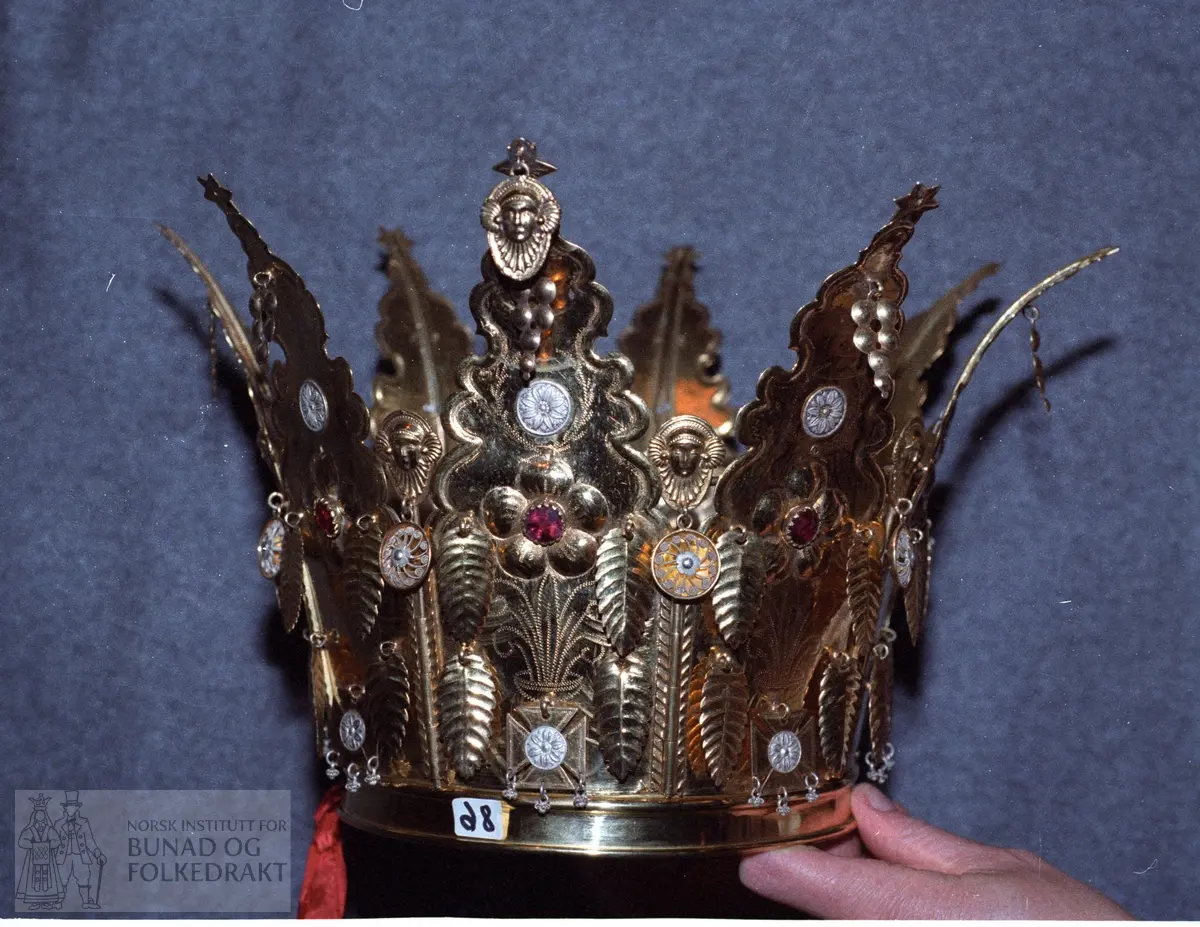 Av forgylt sølv med utpressa mønster. Krona er samansett av 8 delar som er felt ned i ein sarg. Dei 8 delane er haldne saman av ein pinne som blir tredd gjennom pipeforma hol (som dør/vundushekte), sjå foto. Pinnen har andletforma topp samt heng med filigransrose, diameter 2,2 cm. Kvar hovuddel har ei rose av fem kuvne rundingar med raud stein i midten. Gravert blad og stilkmønster under og over rosa. Under mønstret er det eit krossforma heng med innfelt fembladrose - runding i oksydert sylv. Ein litt større fembladroserunding er sett over rosa. Øvst har krona andletheng i kvar del samt fire bladforma heng og eit dråpeforma heng av sju rundingar.