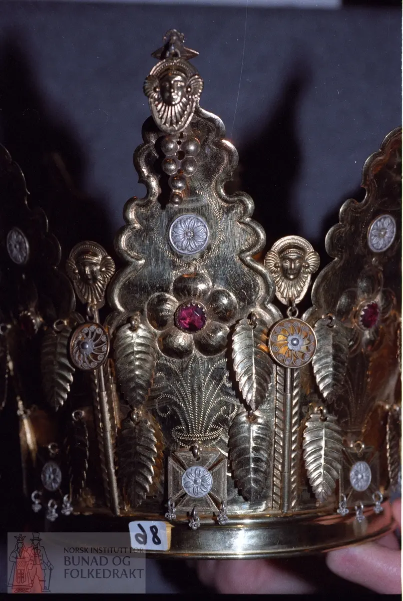 Av forgylt sølv med utpressa mønster. Krona er samansett av 8 delar som er felt ned i ein sarg. Dei 8 delane er haldne saman av ein pinne som blir tredd gjennom pipeforma hol (som dør/vundushekte), sjå foto. Pinnen har andletforma topp samt heng med filigransrose, diameter 2,2 cm. Kvar hovuddel har ei rose av fem kuvne rundingar med raud stein i midten. Gravert blad og stilkmønster under og over rosa. Under mønstret er det eit krossforma heng med innfelt fembladrose - runding i oksydert sylv. Ein litt større fembladroserunding er sett over rosa. Øvst har krona andletheng i kvar del samt fire bladforma heng og eit dråpeforma heng av sju rundingar.