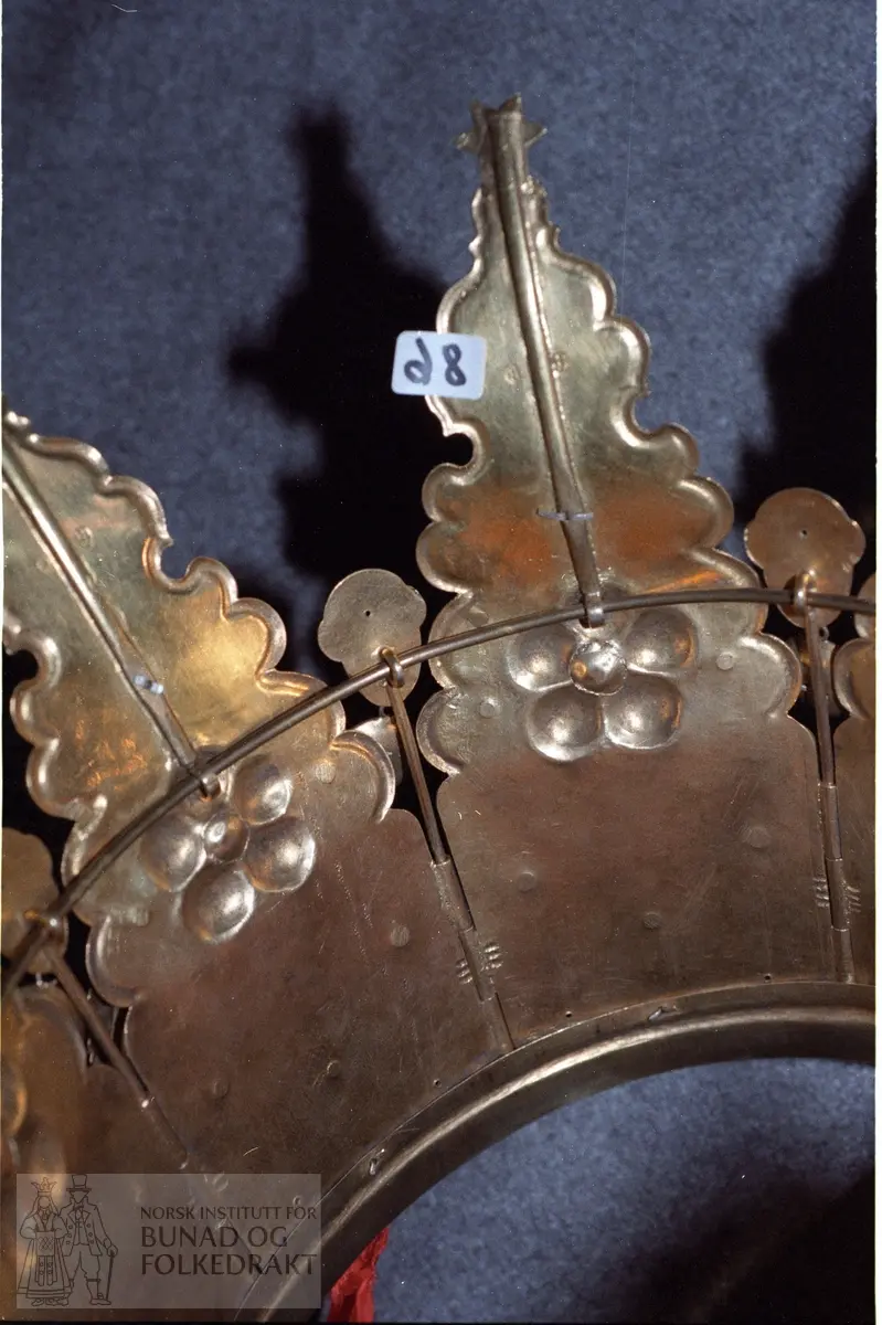 Av forgylt sølv med utpressa mønster. Krona er samansett av 8 delar som er felt ned i ein sarg. Dei 8 delane er haldne saman av ein pinne som blir tredd gjennom pipeforma hol (som dør/vundushekte), sjå foto. Pinnen har andletforma topp samt heng med filigransrose, diameter 2,2 cm. Kvar hovuddel har ei rose av fem kuvne rundingar med raud stein i midten. Gravert blad og stilkmønster under og over rosa. Under mønstret er det eit krossforma heng med innfelt fembladrose - runding i oksydert sylv. Ein litt større fembladroserunding er sett over rosa. Øvst har krona andletheng i kvar del samt fire bladforma heng og eit dråpeforma heng av sju rundingar.