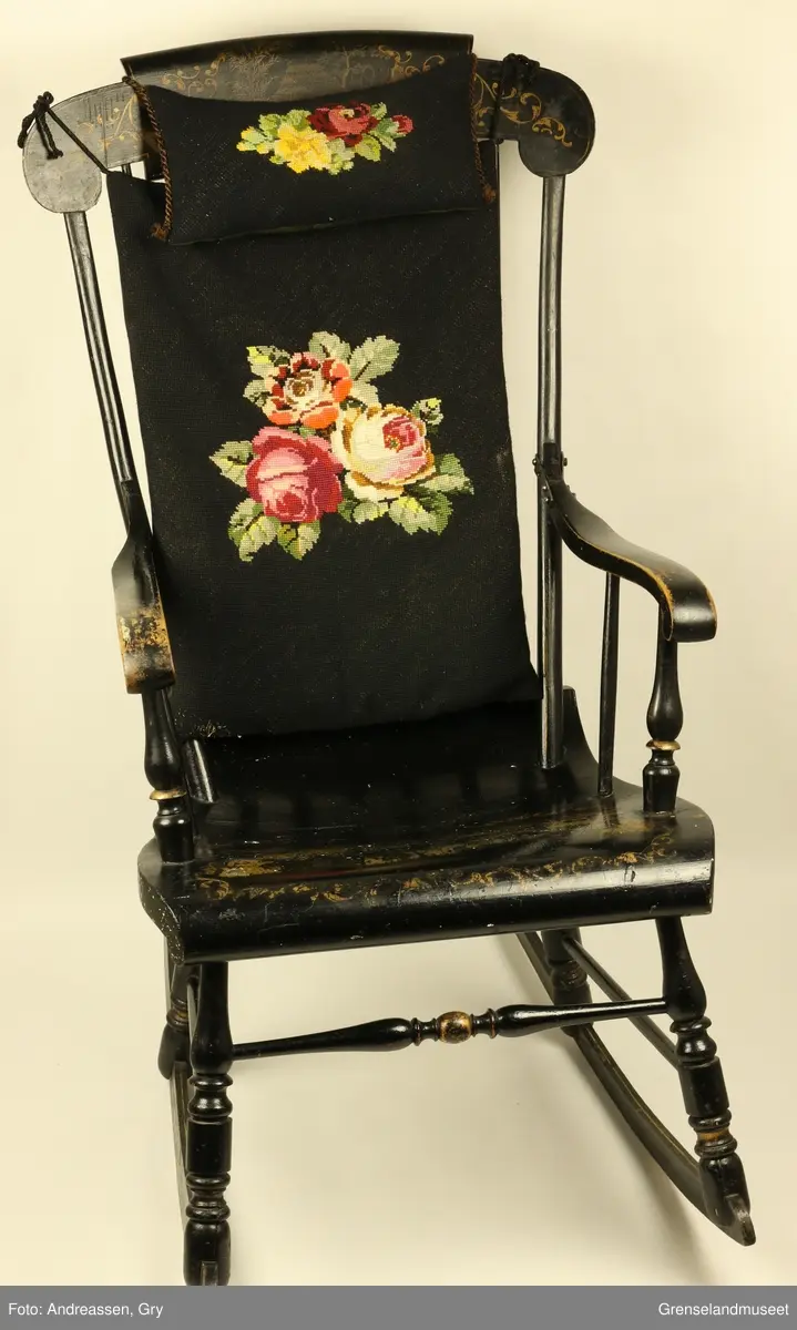 Nakkeputen og ryggputen er i svart aida med brodert rosemønster. Gyngestolen har gulltegninger på setet og rygg.