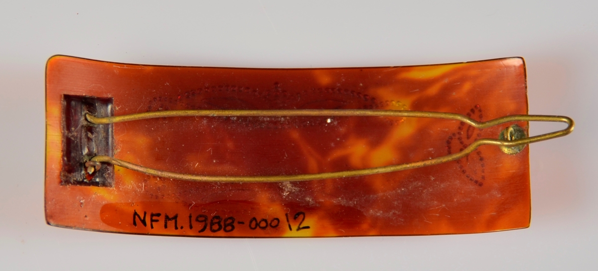 Hårspenne i brunt, marmorert, gjennomskineleg materiale med sølvfarga dekor og metall klype på bakside.