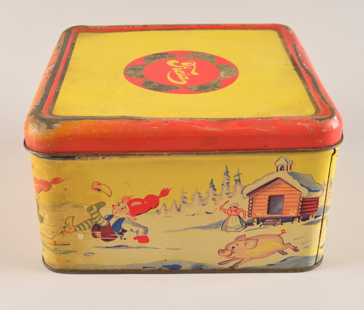 Boks med hengsla lok. Jarnblikk. Trykk på sidene på gul bakgrunn. Motiv er nisser på slede dradd av griser.