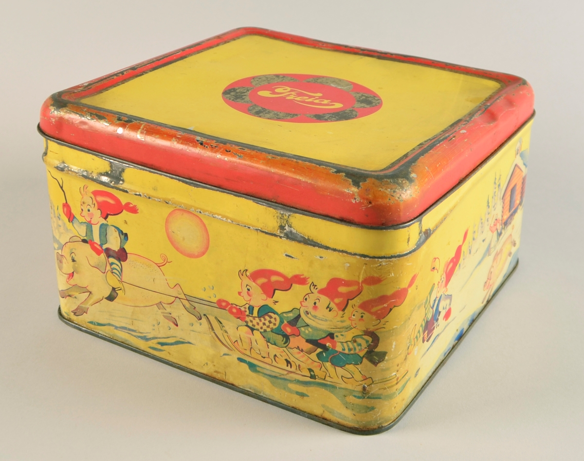 Boks med hengsla lok. Jarnblikk. Trykk på sidene på gul bakgrunn. Motiv er nisser på slede dradd av griser.