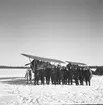 Gruppfoto av flygare från F 19 i Märkäjärvi vid Svenska frivilligkåren i Finland, F 19. I bakgrunden står två flygplan.