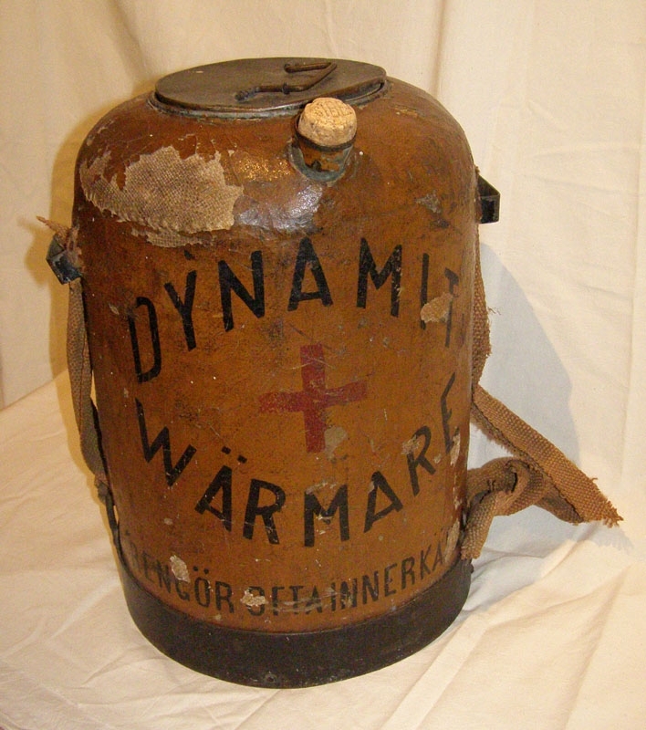 Dynamitvärmare med behållare klädd med brunmålad säckväv. 
Lock av metall och kork. Pip i metall att hälla ur. Bärhandtag i säckväv.
Text i svart: "Nitroglycerinbolaget. Stockholm. Med såpa och varmt vatten. Dynamit-värmare. Rengör ofta innerkärlet".