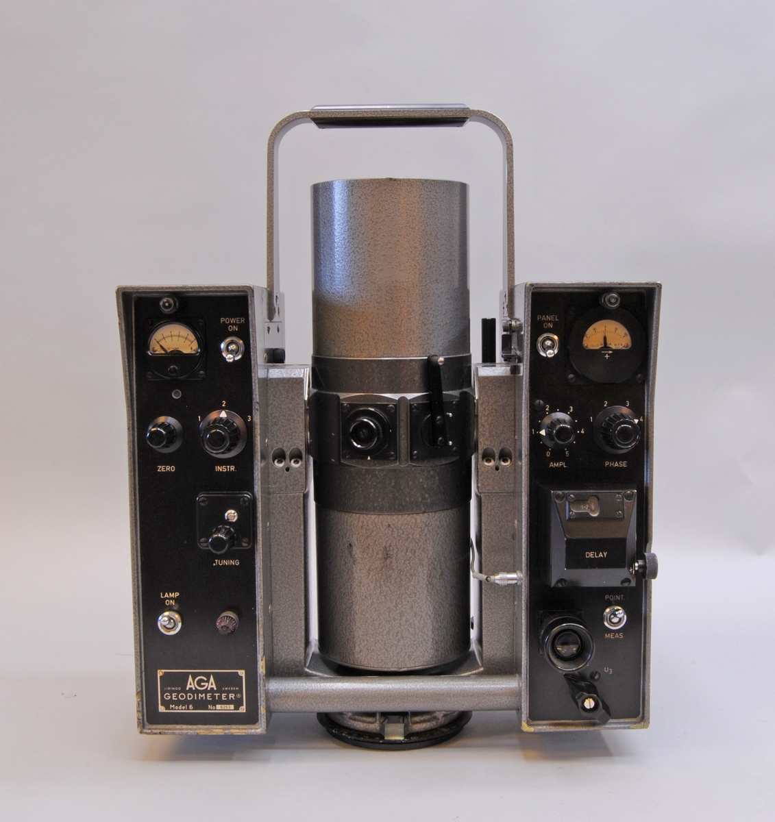 AGA Geodimeter modell 6. Mätinstrumentet har ett grått plåthölje. I mitten av instrumentet finns en cylinderformad tub som går att vinkla till horisontellt läge och skruvas åt för att fastsättas. På sidorna om tuben är två rektangulärt stående partier med olika reglage på den sidan som instrumentet manövreras från. Instrumentet står på en rund vridbar platta som är avsedd att fästas i ett stativ. Se även fodral Jvm23132:2-3 samt tillbehör Jvm23132:4-9.