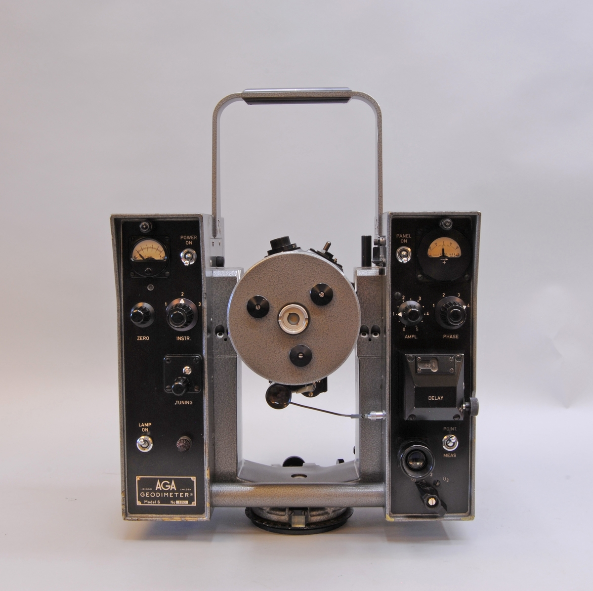 AGA Geodimeter modell 6. Mätinstrumentet har ett grått plåthölje. I mitten av instrumentet finns en cylinderformad tub som går att vinkla till horisontellt läge och skruvas åt för att fastsättas. På sidorna om tuben är två rektangulärt stående partier med olika reglage på den sidan som instrumentet manövreras från. Instrumentet står på en rund vridbar platta som är avsedd att fästas i ett stativ. Se även fodral Jvm23132:2-3 samt tillbehör Jvm23132:4-9.