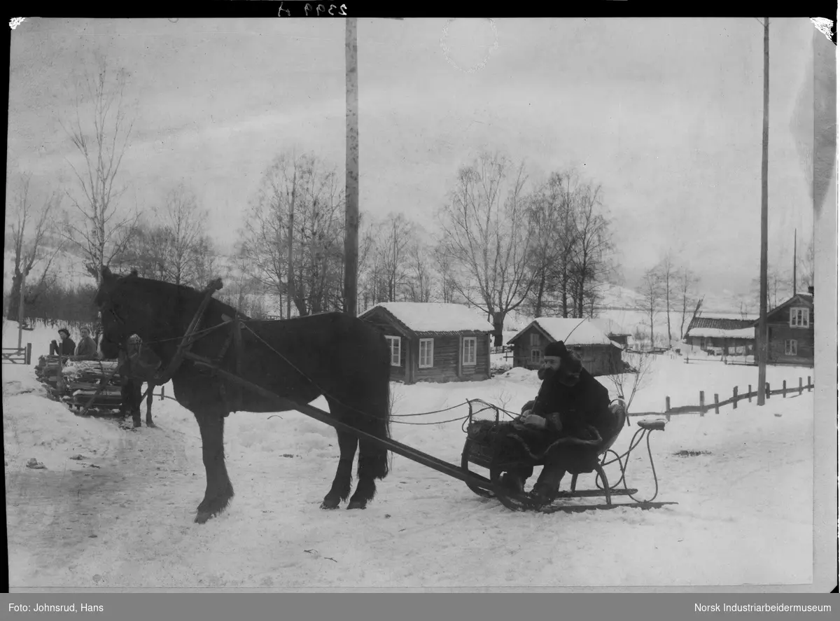 Doktor Helmers på vei til sykebesøk med hest og slede. Hest med slede, to mennesker og trevirke i bakgrunnen. Laftede bygninger, kvinne stående på tunet, klesvask henger til tørk.