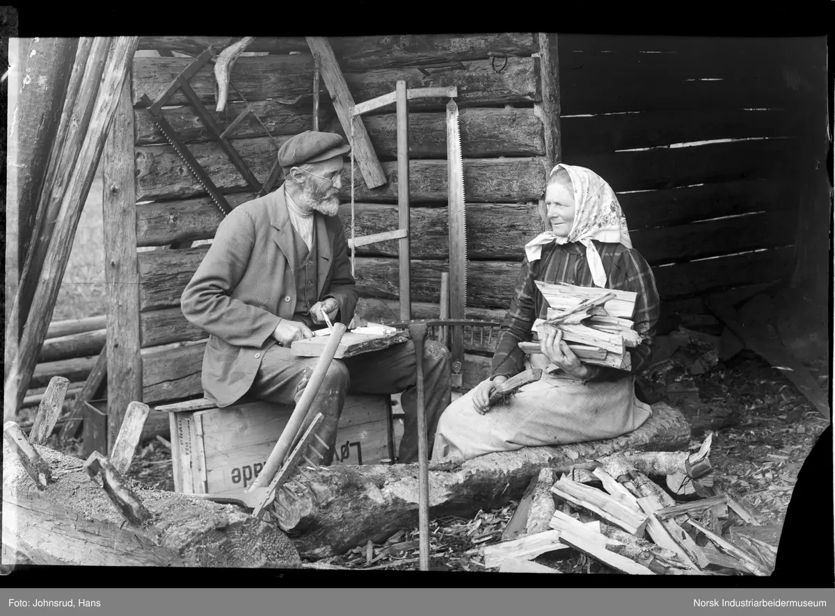 Ole og Berthe Marie Johnsrud sittende utenfor laftet vedskjul. Kvinnen med ved i armene, mann i arbeid med å spikke rivetenner.
