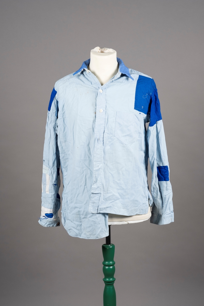 En lyselå skjorte reparert med påsydde lapper i andre blåfarger. Skjorten har tre knapper i plast, mangler to knapper. Det er brystlomme på høyre side og knapp på venstre mansjett. Fangenummer er påført under kragen på ryggen. Det kan være brodert en liten fisk over brystlommen.