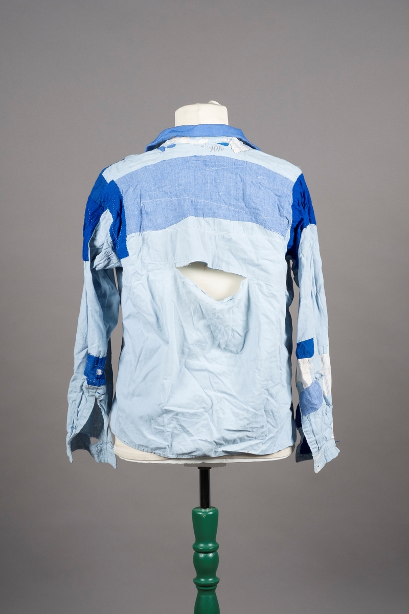 En lyselå skjorte reparert med påsydde lapper i andre blåfarger. Skjorten har tre knapper i plast, mangler to knapper. Det er brystlomme på høyre side og knapp på venstre mansjett. Fangenummer er påført under kragen på ryggen. Det kan være brodert en liten fisk over brystlommen.