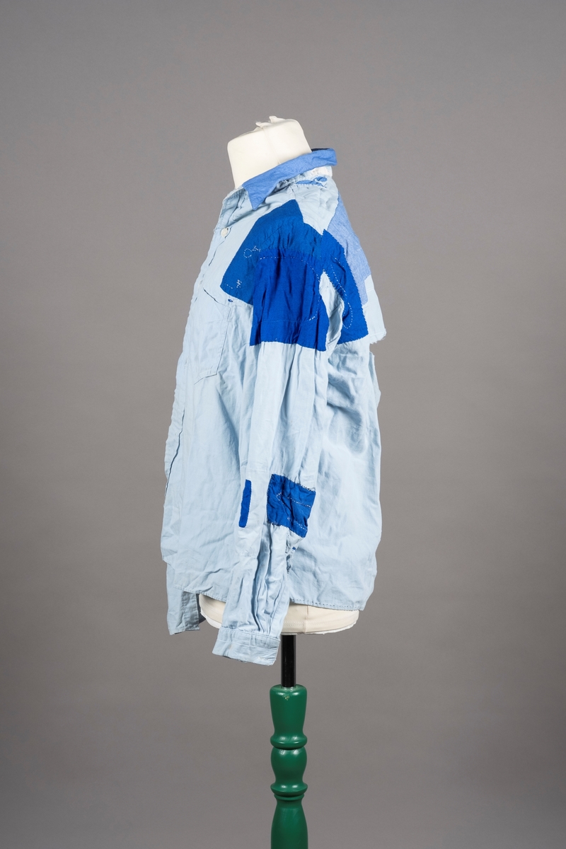 En lyselå skjorte reparert med påsydde lapper i andre blåfarger. Skjorten har tre knapper i plast, mangler to knapper. Det er brystlomme på høyre side og knapp på venstre mansjett. Fangenummer er påført under kragen på ryggen. Det kan være brodert en liten fisk over brystlommen.