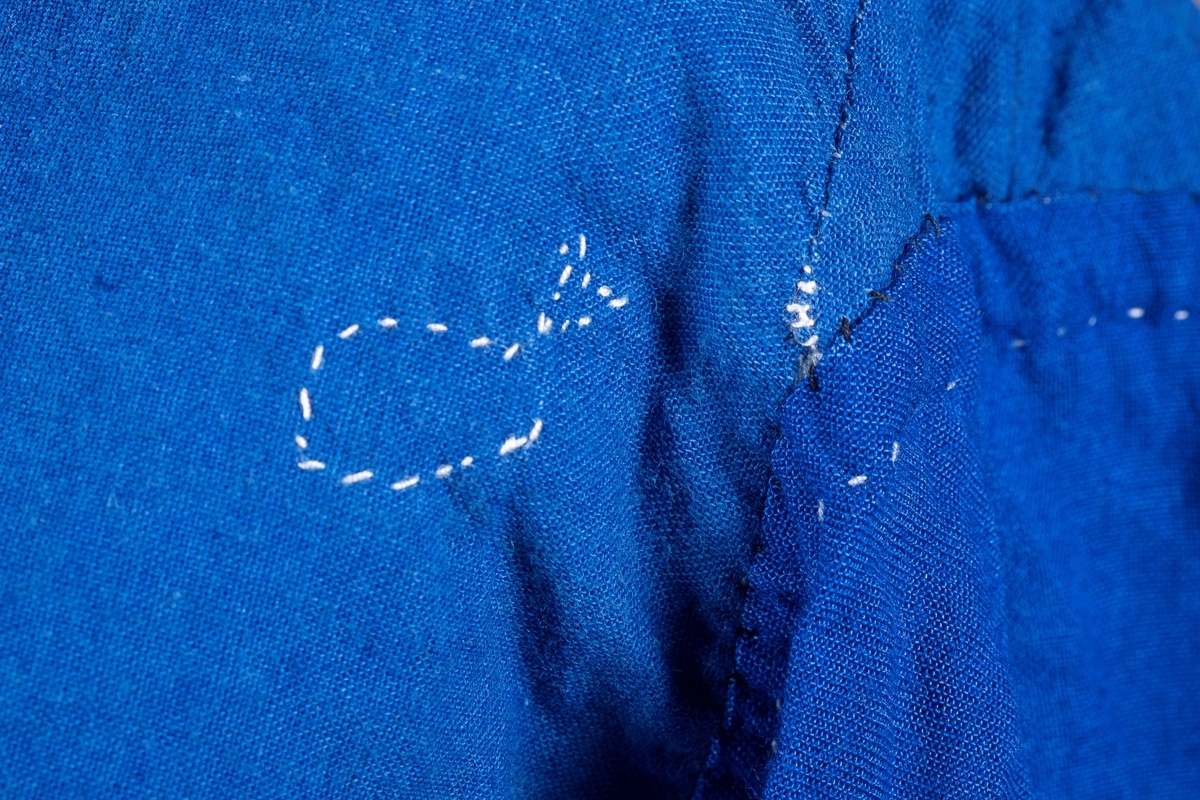 En lyselå skjorte reparert med påsydde lapper i andre blåfarger. Skjorten har tre knapper i plast, mangler to knapper. Det er brystlomme på høyre side og knapp på venstre mansjett. Fangenummer er påført under kragen på ryggen. Det kan være brodert en liten fisk over brystlommen.