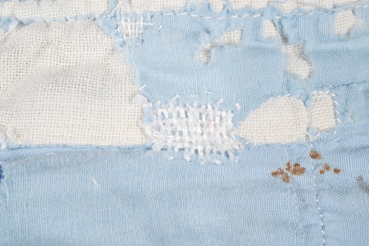 En lyselå skjorte reparert med påsydde lapper i andre blåfarger. Skjorten har tre knapper i plast, mangler to knapper. Det er brystlomme på høyre side og knapp på venstre mansjett. Fangenummer er påført under kragen på ryggen. Det kan være brodert en liten fisk over brystlommen.