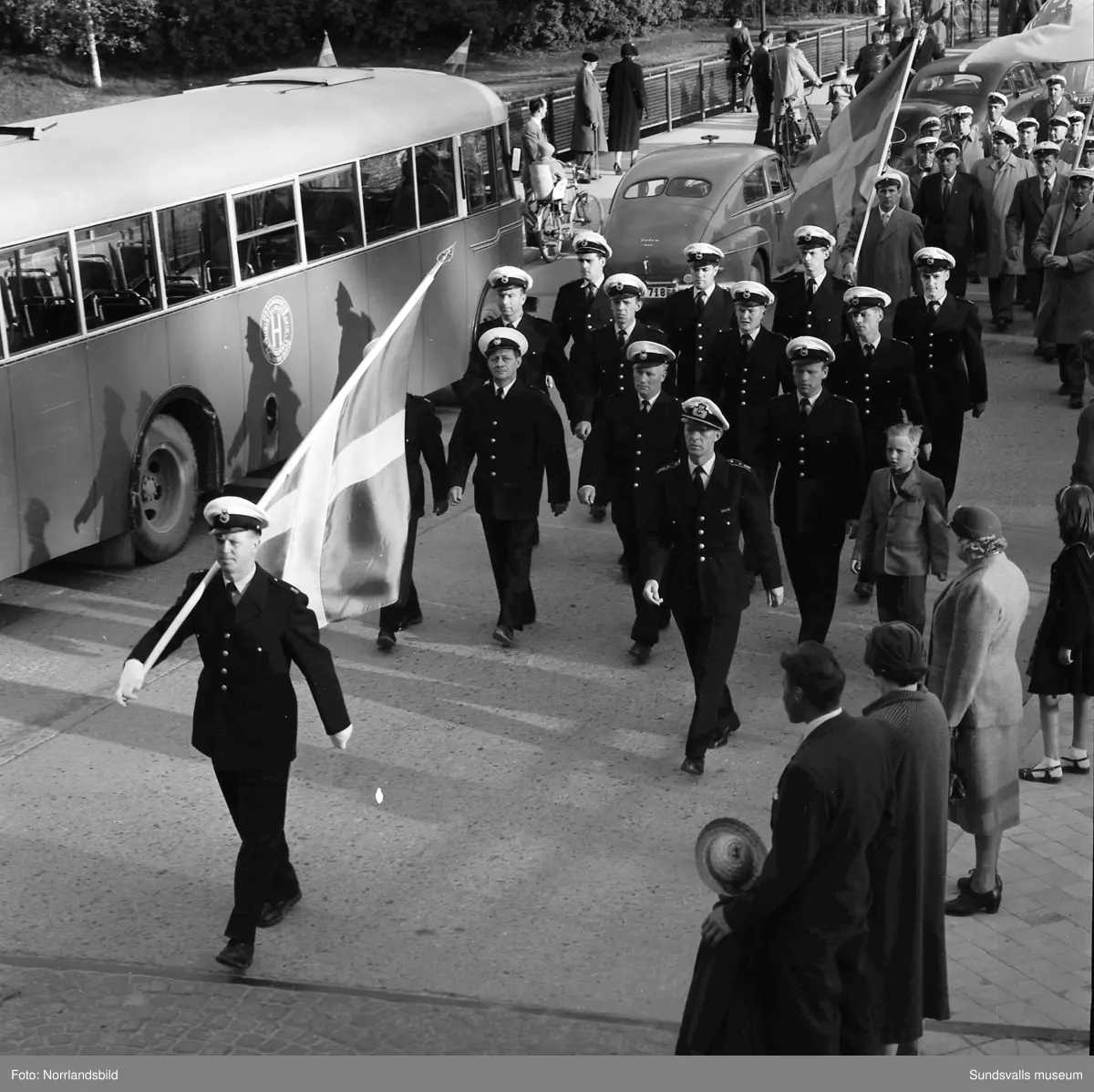 En stor serie bilder från firandet av Svenska flaggans dag 1956. Parad på Storbron med paradorkester, militär, scouter och mängder av olika föreningar och förbund. Därefter högtidlig uppställning i Idrottsparken med bland annat tal, körsång, gymnastikuppvisning och folkmusik.