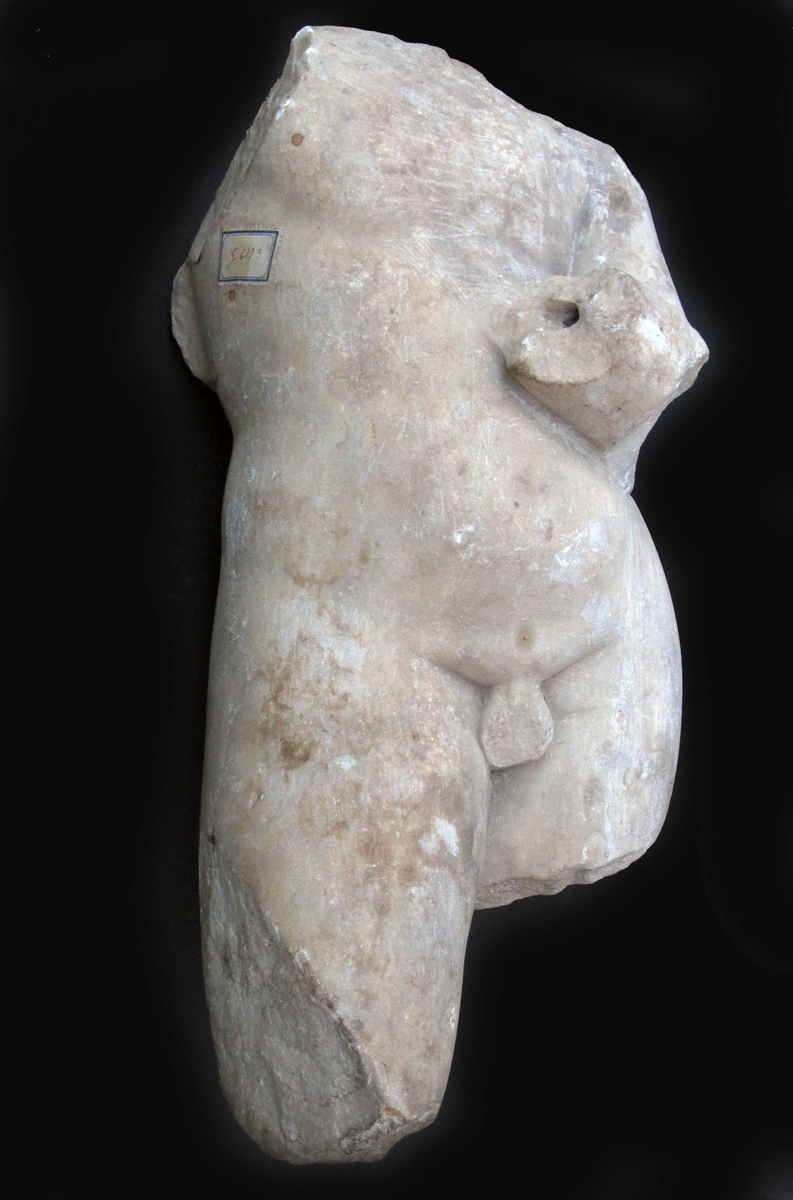Torso av buespennende Eros, romersk keisertids kopi av gresk original.  Fin krystalinsk marmor. Figuren fragmentarisk bevart; hodet, høyre arm, vifigene og  bena delvis avslått. Figuren bøyer venstre arm opp mot brystet,  armen brukket av like over håndleddet. På bruddflaten et dypt boret hull, etter nagle som har holdt hånden fast. Figuren står på det venstre benet,  det høyre er skjøvet frem, og antageligvis har  kneet vært bøyet. Figuren knekker i hoften, og  dette gir den en s-formet bevegelse som går gjennom kroppen. Skulpturen er en romersk keisertidskopi av  den greske billedhugger Lysippos verk.