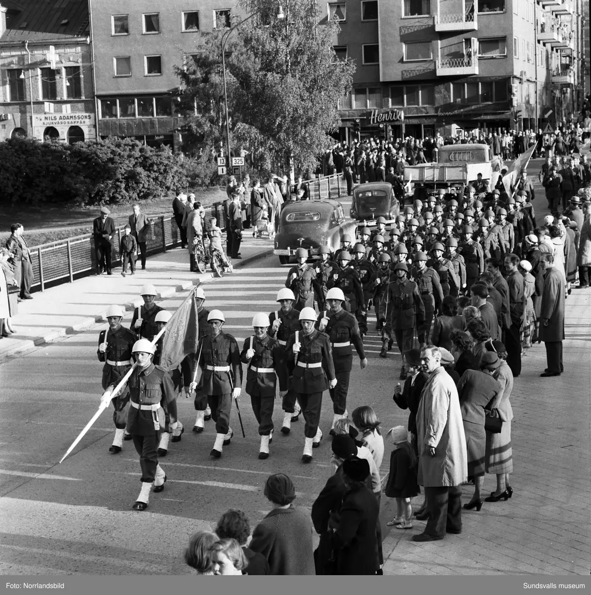 En stor serie bilder från firandet av Svenska flaggans dag 1956. Parad på Storbron med paradorkester, militär, scouter och mängder av olika föreningar och förbund. Därefter högtidlig uppställning i Idrottsparken med bland annat tal, körsång, gymnastikuppvisning och folkmusik.