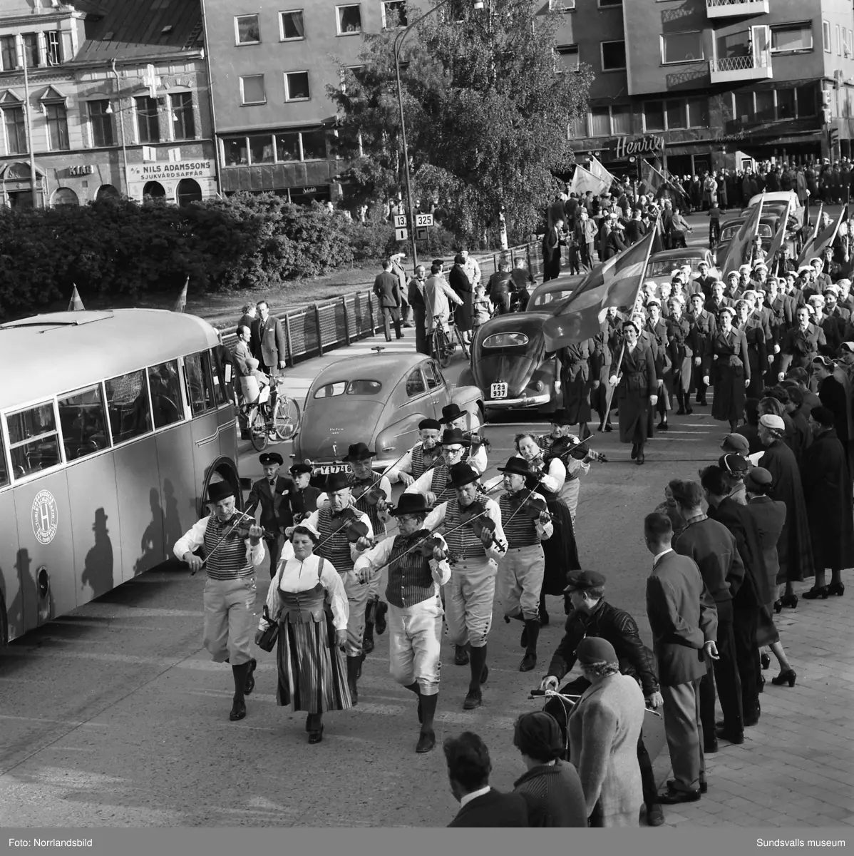 En stor serie bilder från firandet av Svenska flaggans dag 1956. Parad på Storbron med paradorkester, militär, scouter och mängder av olika föreningar och förbund. Därefter högtidlig uppställning i Idrottsparken med bland annat tal, körsång, gymnastikuppvisning och folkmusik.