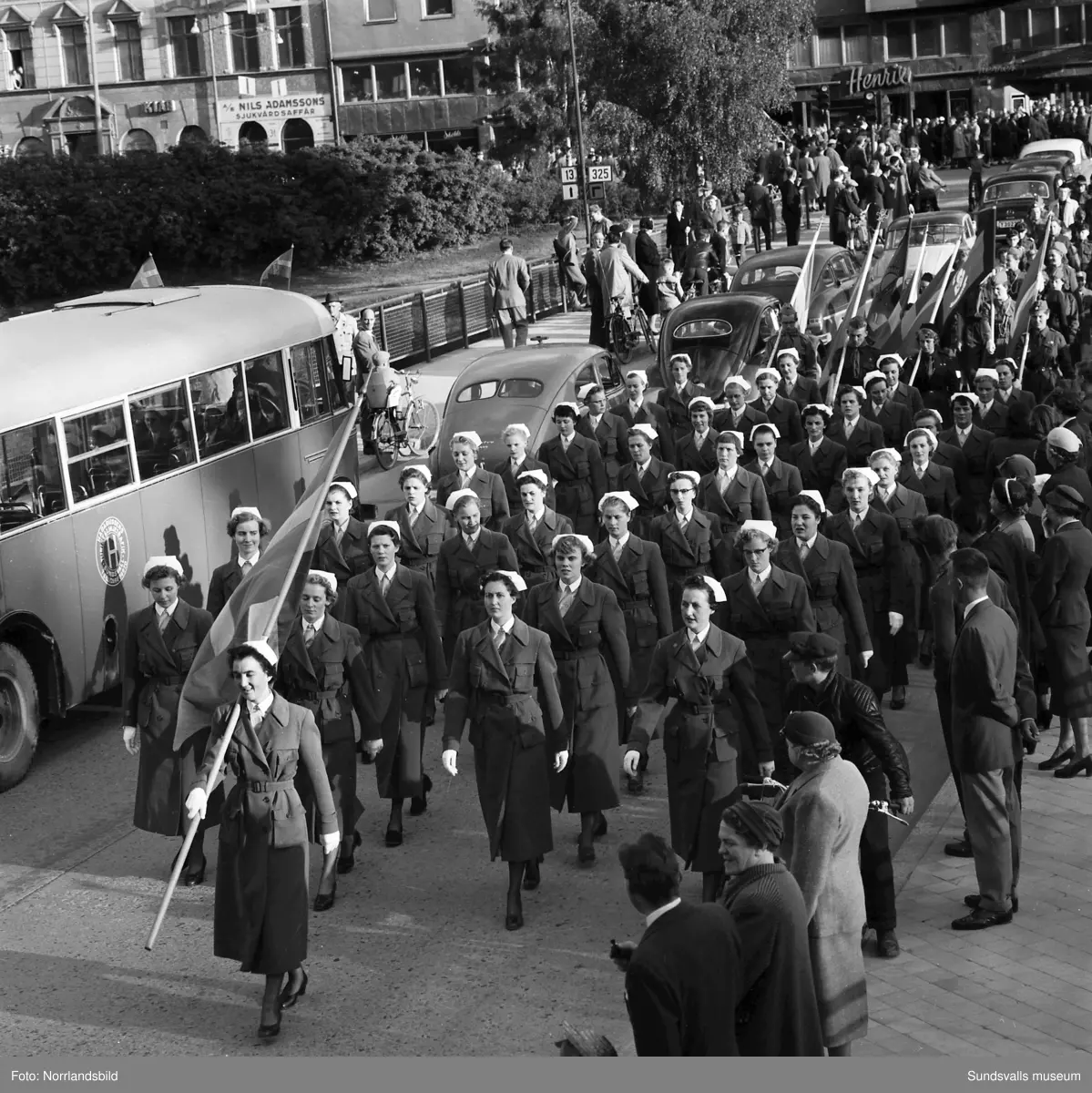 En stor serie bilder från firandet av Svenska flaggans dag 1956. Parad på Storbron med paradorkester, militär, scouter och mängder av olika föreningar och förbund. Därefter högtidlig uppställning i Idrottsparken med bland annat tal, körsång, gymnastikuppvisning och folkmusik.