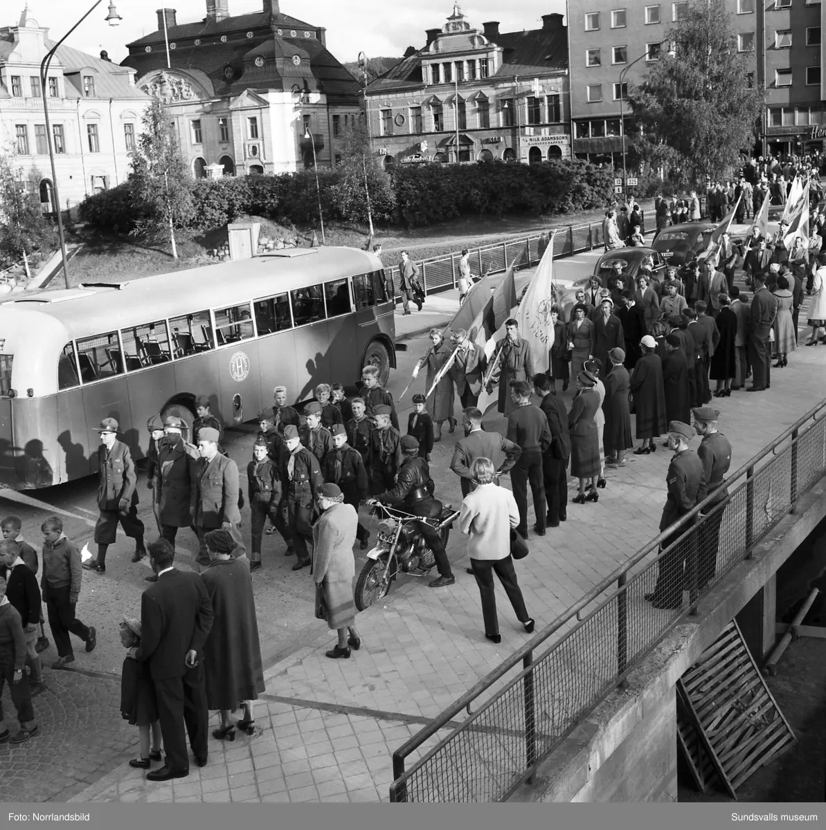 En stor serie bilder från firandet av Svenska flaggans dag 1956. Parad på Storbron med paradorkester, militär, scouter och mängder av olika föreningar och förbund. Därefter högtidlig uppställning i Idrottsparken med bland annat tal, körsång, gymnastikuppvisning och folkmusik.