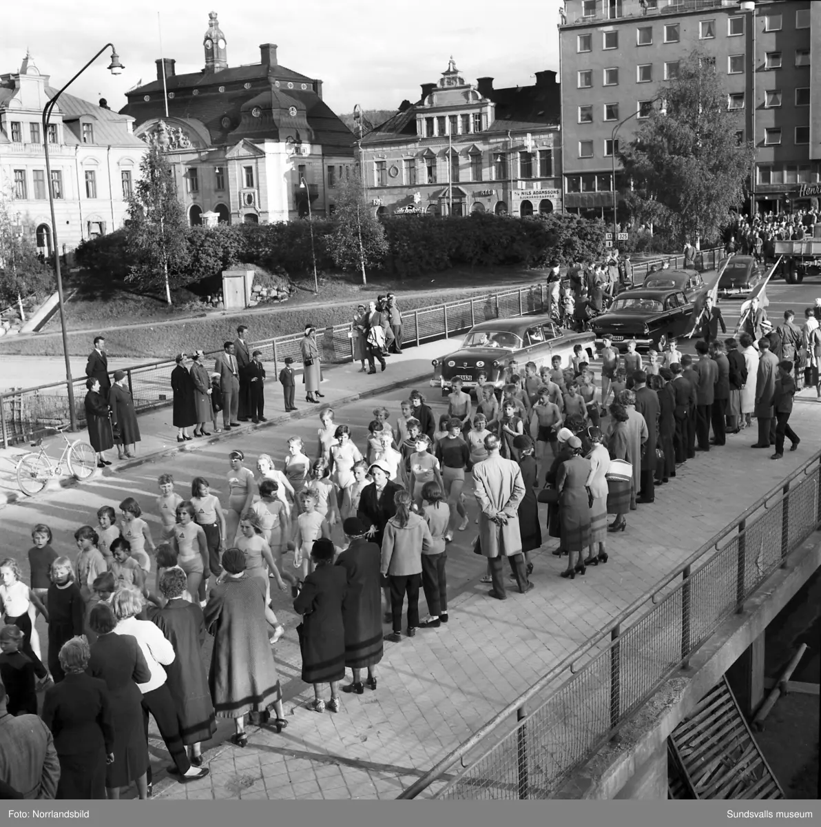 En stor serie bilder från firandet av Svenska flaggans dag 1956. Parad på Storbron med paradorkester, militär, scouter och mängder av olika föreningar och förbund. Därefter högtidlig uppställning i Idrottsparken med bland annat tal, körsång, gymnastikuppvisning och folkmusik.
