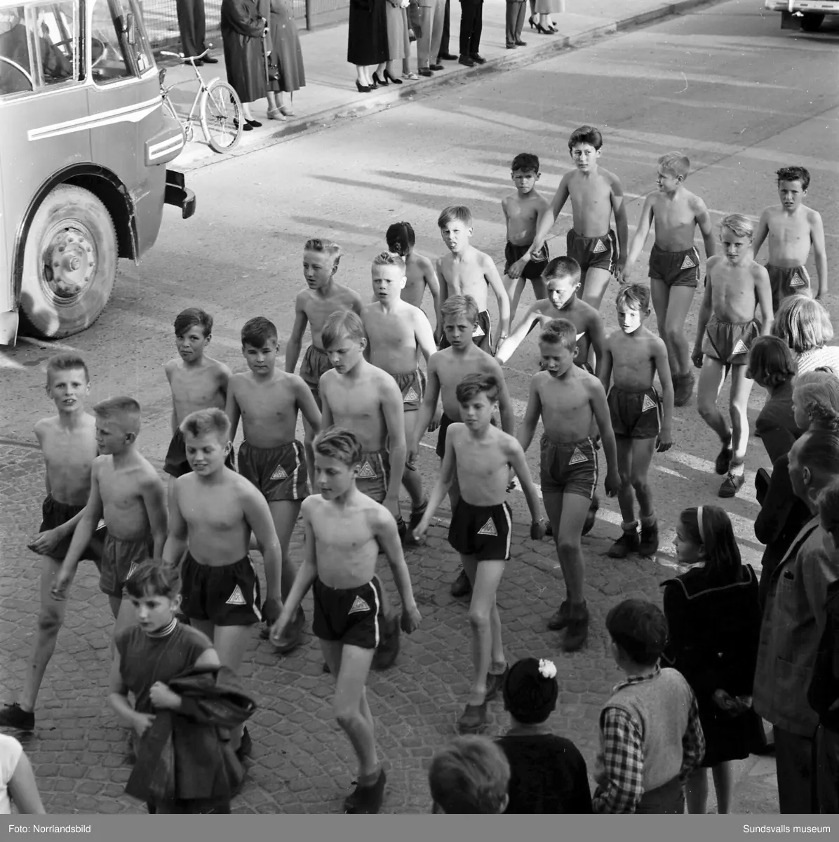 En stor serie bilder från firandet av Svenska flaggans dag 1956. Parad på Storbron med paradorkester, militär, scouter och mängder av olika föreningar och förbund. Därefter högtidlig uppställning i Idrottsparken med bland annat tal, körsång, gymnastikuppvisning och folkmusik.
