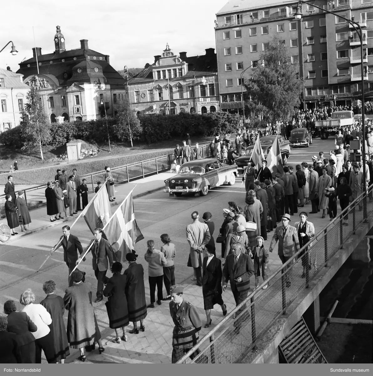 En stor serie bilder från firandet av Svenska flaggans dag 1956. Parad på Storbron med paradorkester, militär, scouter och mängder av olika föreningar och förbund. Därefter högtidlig uppställning i Idrottsparken med bland annat tal, körsång, gymnastikuppvisning och folkmusik.