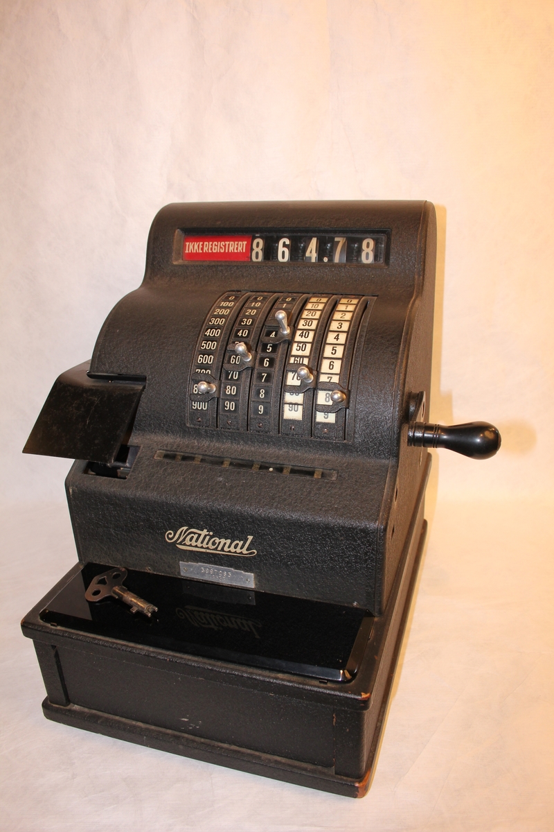 Kassa-apparat med sokkel, pengeskuff og sveiv, 5 skalaer for kr. og øre. Vindauga for kronesum framme og bak. 5 knottar. Nøkkel til skuff.