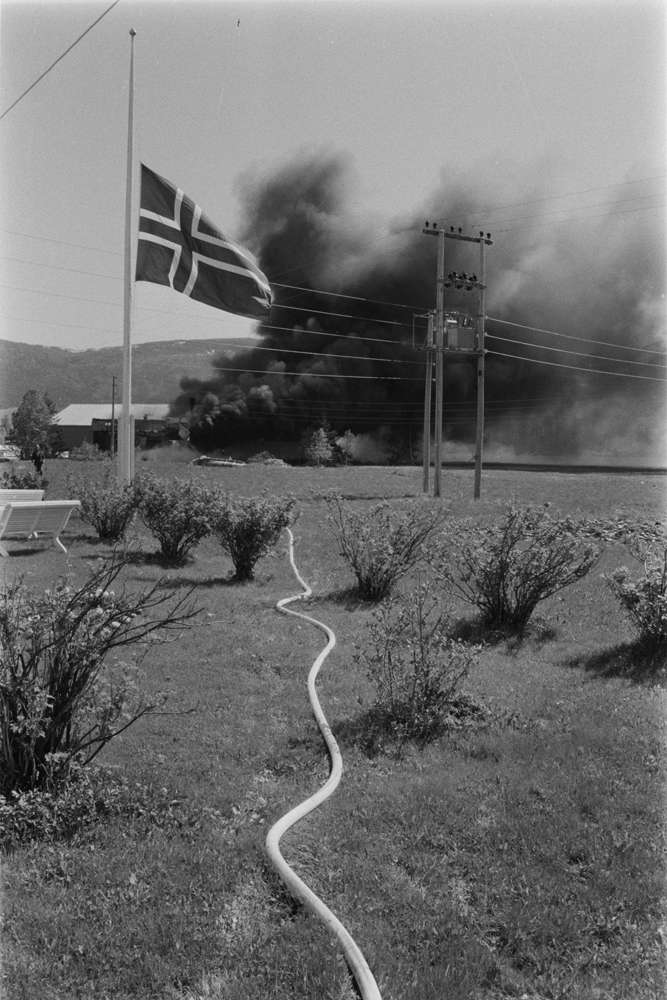 Båtfabrikken Polar-Industri A/S i Drevvatnet totalskadd ved brann 16 Juni 1978. Fabrikken brenner, mye røyk.
Brannslange over plen, flagg på halv stang.