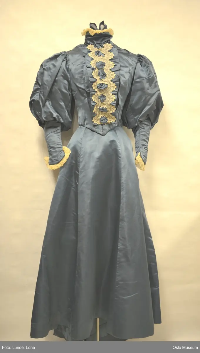 Dueblå kjole i silkeduchesse. Ecrufarget blonde foran på liv og rynket chiffon i erme og hals. Store skinkeermer.