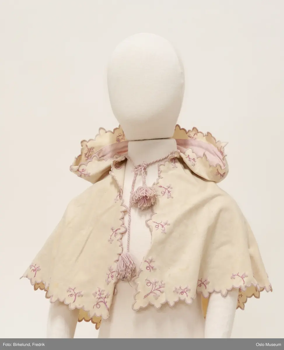 Barnecape brodert med tunger i kanten og blomster i to farger. 
Liten sløyfe på hodet i stoffet, snor med pomponger i rosa ullgarn
Størrelse: 1/2- 1 år