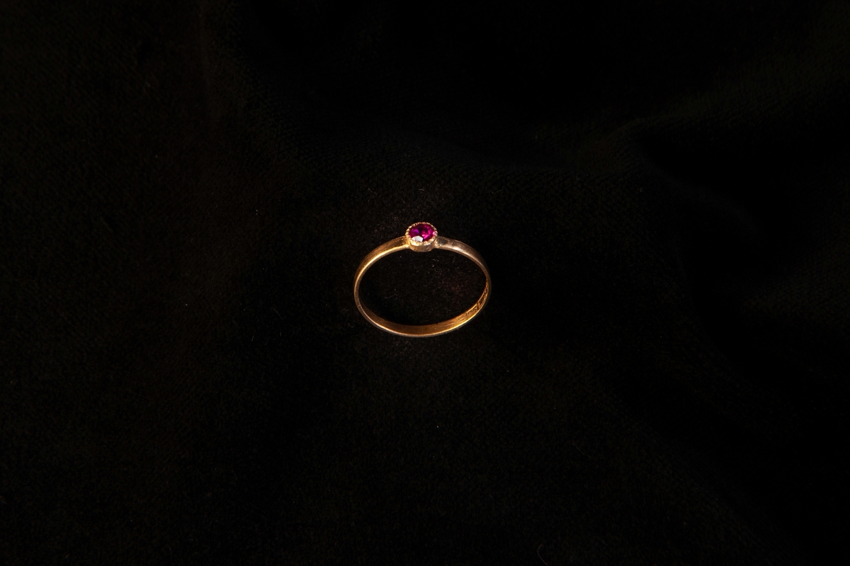 Smal guldring med infattad ljusröd sten (troligen simili (glas), möjligen rubin(?). En tillhörande rund smyckesask i papp.