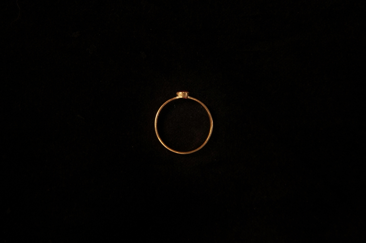 Smal guldring med infattad ljusröd sten (troligen simili (glas), möjligen rubin(?). En tillhörande rund smyckesask i papp.