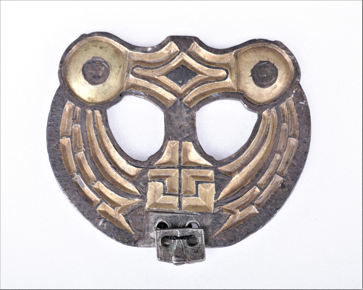 Sølvbeslag og en liten del av dens festeinnretning, forgylt med nielloinnlegg, som type R. 200 d fra eldre jernalder. Utsmykning til sverdslire. Funnet i en gravhaug på gården Mellom-Kvem.