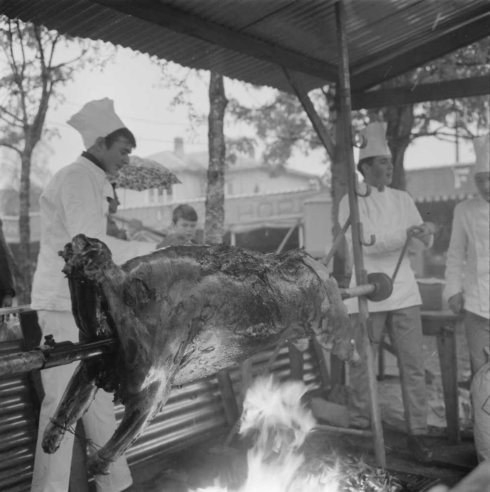 Tiendebyttet 1966. Grilling av hel gris over åpen flamme under blikkskur. Menn med kokkeluer.