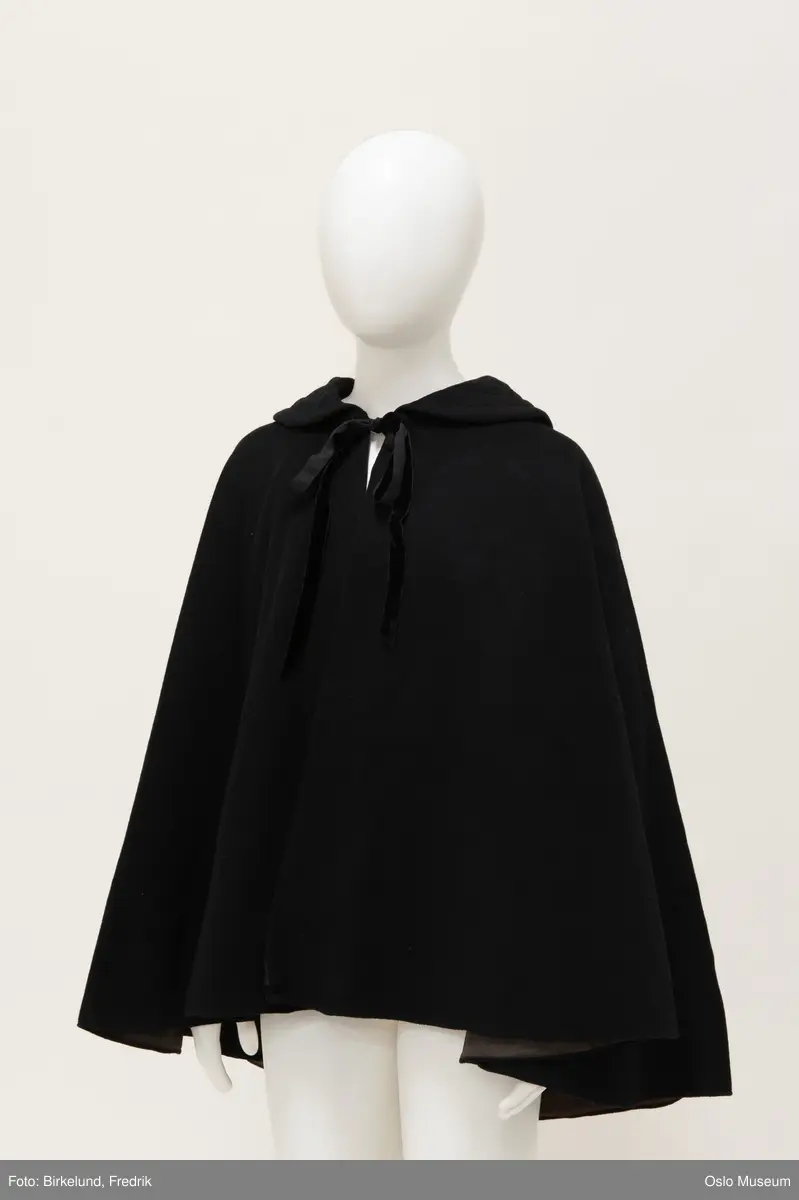 Sort, kort cape. Lukkes med hekte og knytebånd i halsen.