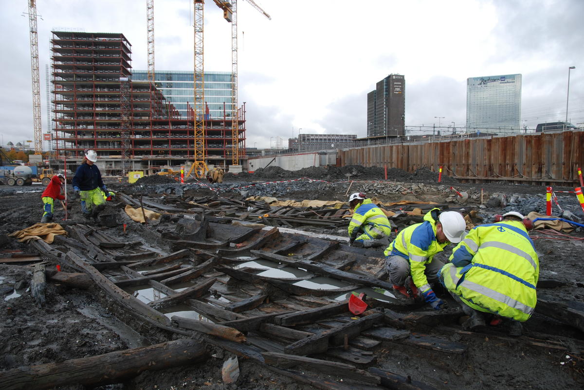 Tre arkeologer i gult arbeidstøy og med hvite hjelmer sitter på huk og graver i restene av en trebåt som ligger i gjørme. Båten er delvis fylt med vann. En arkeolog i bakgrunnen spar i leire med spade. I bakgrunnen ses Postgirobygget, SAS-hotellet og kjøreledninger til tog over skinnene på Oslo S. Et bygg under oppføring, omgitt av kraner og anleggsmaskiner, står på andre siden av samme tomt som arkeologene arbeider på.