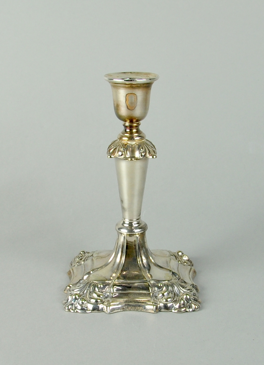 Lysestake av sølv. Lysestaken har firkantet sokkel med rikt profilert dekor. I hjørnene av sokkel er det akantuslignende dekor. På midten av staken er det utgående, akantuslignende dekor.