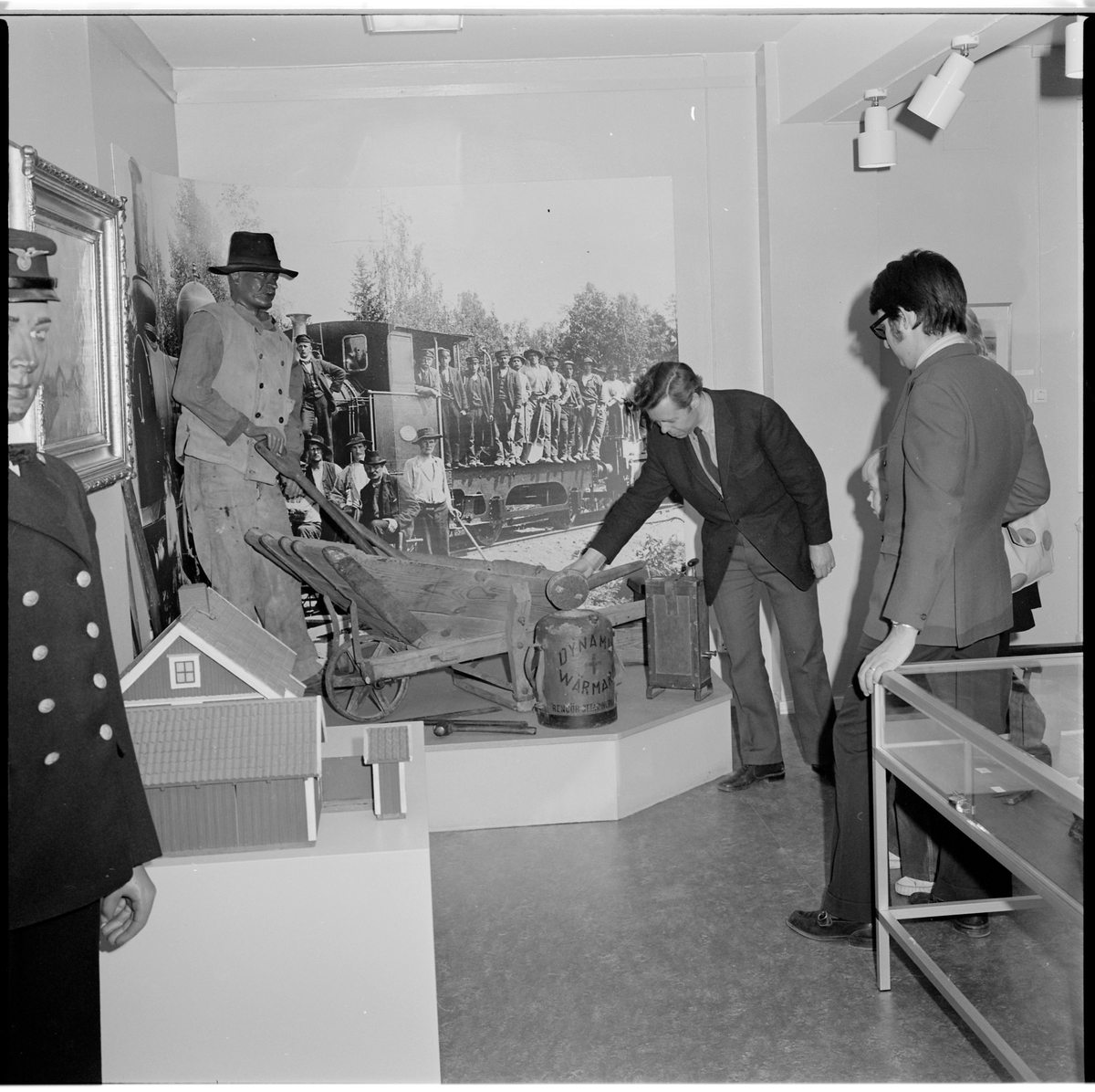 Dynamitvärmare med behållare klädd med brunmålad säckväv. 
Lock av metall och kork. Pip i metall att hälla ur. Bärhandtag i säckväv.
Text i svart: "Nitroglycerinbolaget. Stockholm. Med såpa och varmt vatten. Dynamit-värmare. Rengör ofta innerkärlet".