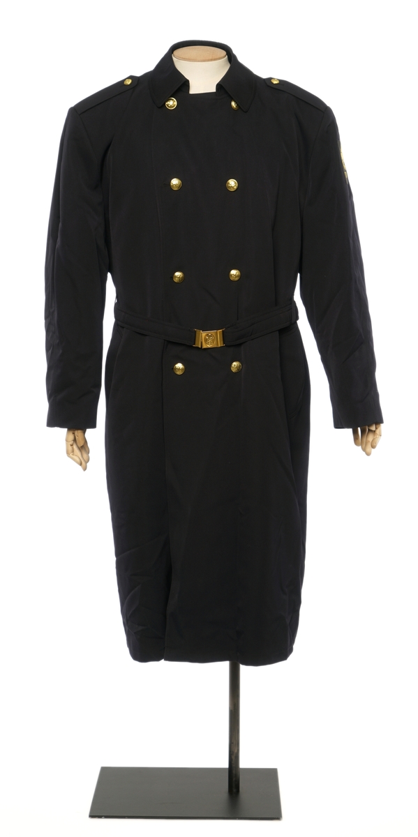 Mørkblå kappe dobbelspent med fire knapper med riksløve-motiv. Epauletter uten tilsatt distinktsjoner. Split i bak, og innebygd belte med sammenlåsende lukking. Avtagbar vattert foder festet av glidelåser rundt inner kanten. Stoffet er finveved kypert blanding av ull og polyester) Jakke er foda med syntetisk sort stoff. Tekstil merke på venstre arm, riksløve med hvit øks omgitt av eikeblader over TOLL.