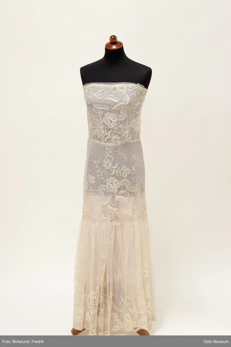 A: Rett fasong fra byste ned over hofte og lårparti. Løsere i skjørtet nedenfor hofte/lår.  Ermeløs med trykknapper i siden. Øvre tøylag er gulhvit blonde med blomsterornamentikk.  Blonden er festet og henger over en rosa underkjole. Det rosa stoffet skinner gjennom og gjør kjolen blek rosa.
B: hårkrans